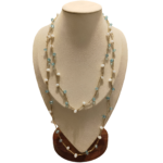 Collana artigianale lunga con catena dorata e pietre in pasta di vetro bianche e celesti, disposta su busto espositivo beige