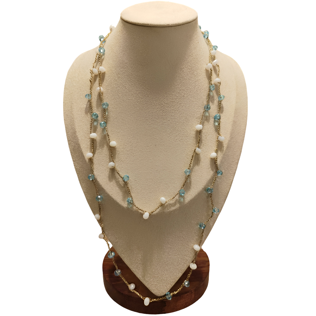 doralie-jewels-collana-design-monopoli-01 Collana artigianale lunga con catena dorata e pietre in pasta di vetro bianche e celesti, disposta su busto espositivo beige